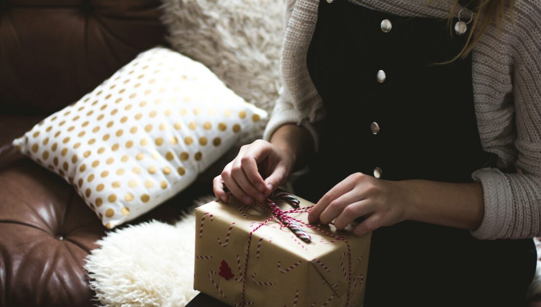 woman in grey sweater and blue denim dungarees sitting on brown and white sofa opening a yellow gift box closeup photography